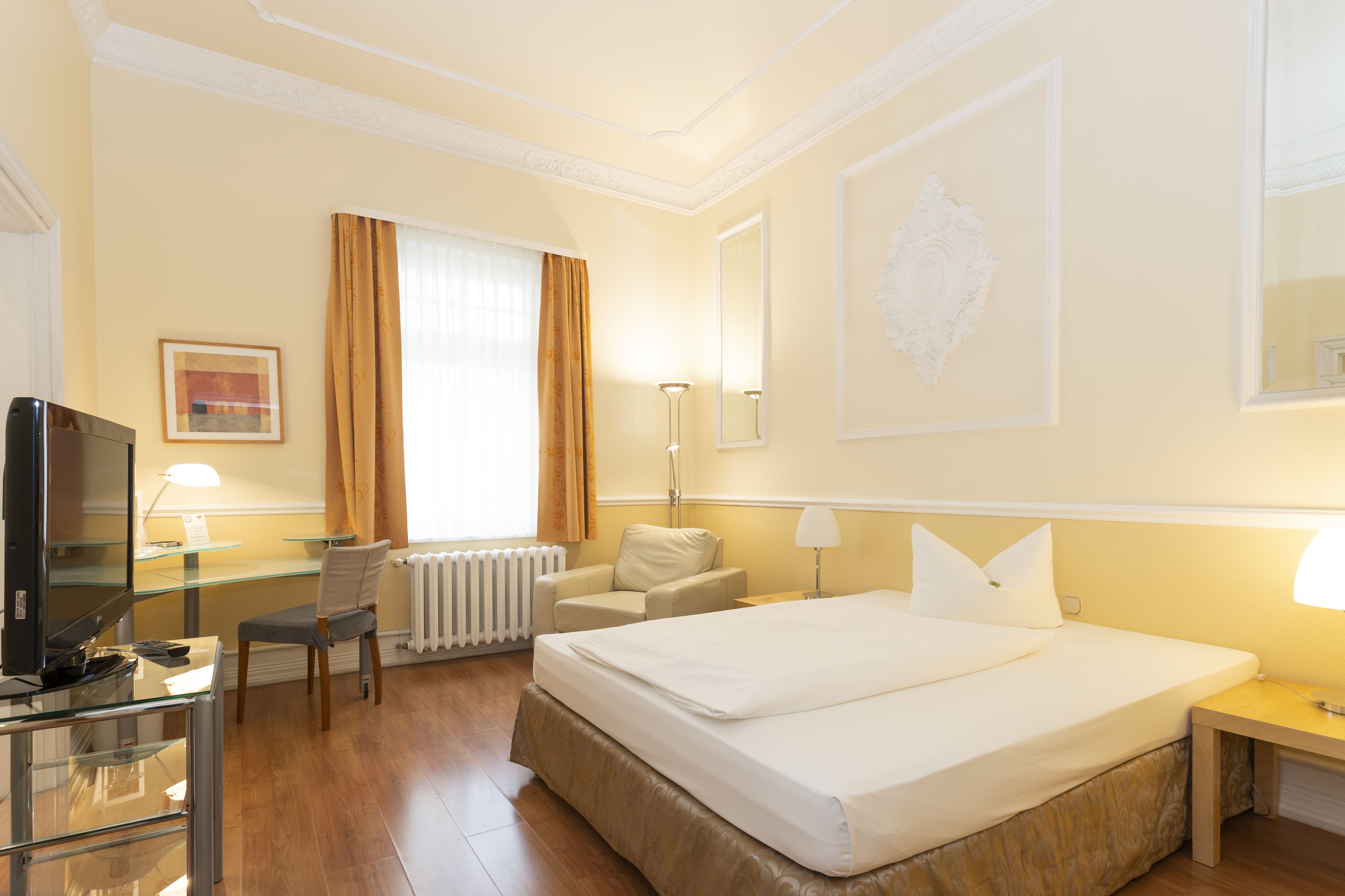 A hotel room with a bed, a desk with a computer, a chair, a radiator, and a window with curtains.