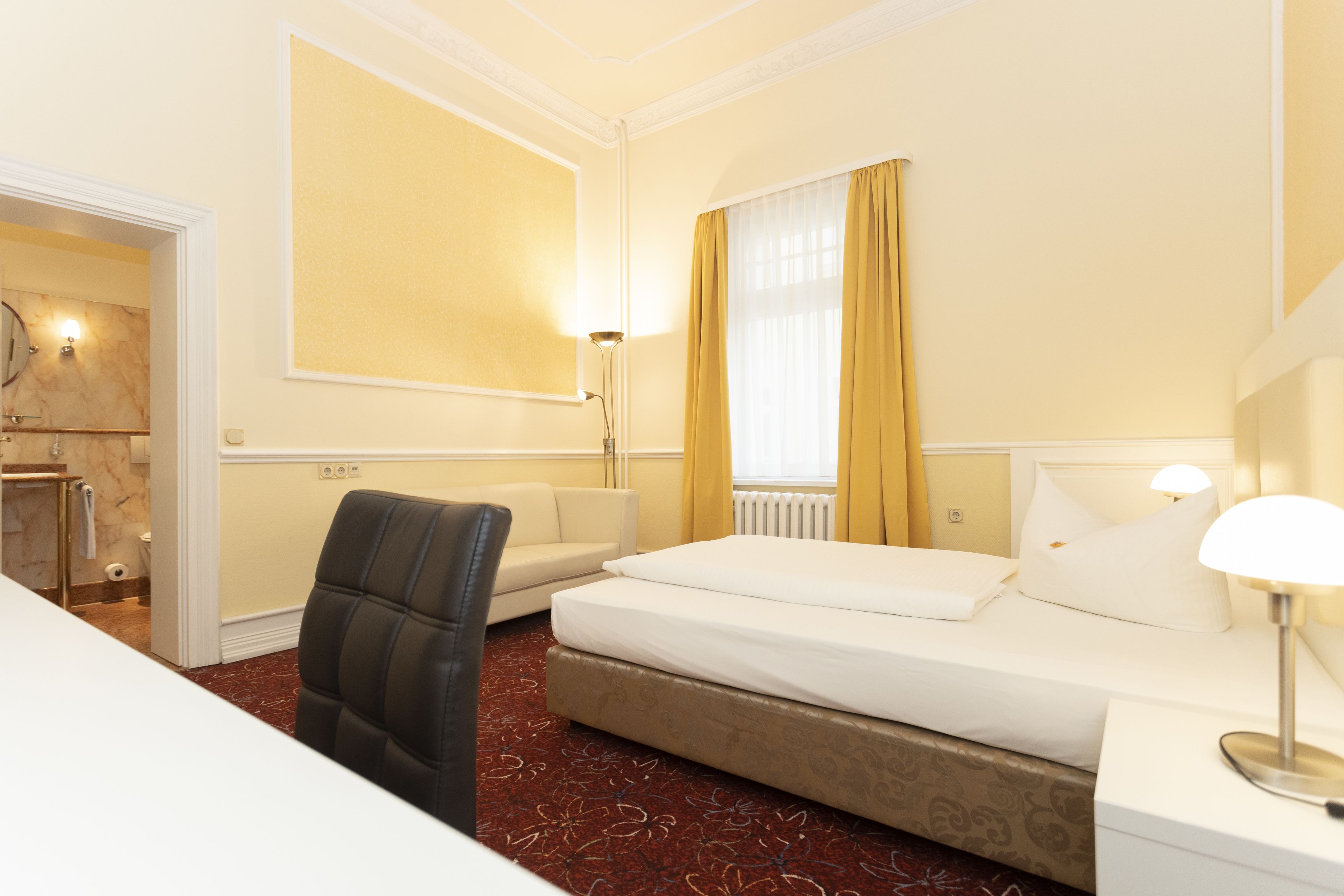 Una camera d'albergo con un letto, una sedia, un comodino, una lampada e una finestra con tende.