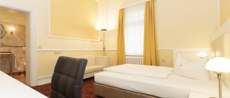 A hotel room with a bed, a chair, a nightstand, a lamp, and a window with curtains.