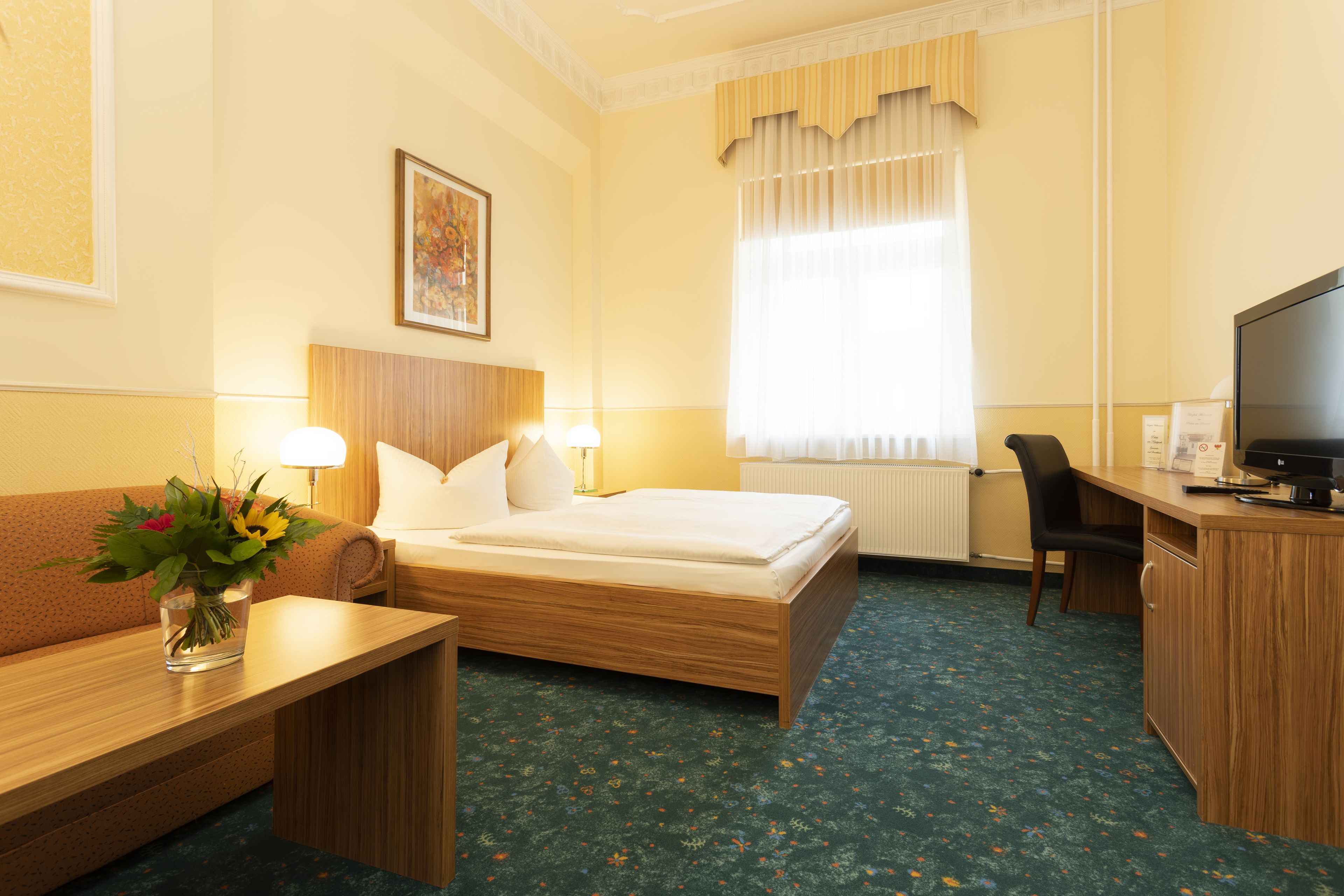 Una habitación de hotel con cama, escritorio, silla, televisor y un jarrón con flores.