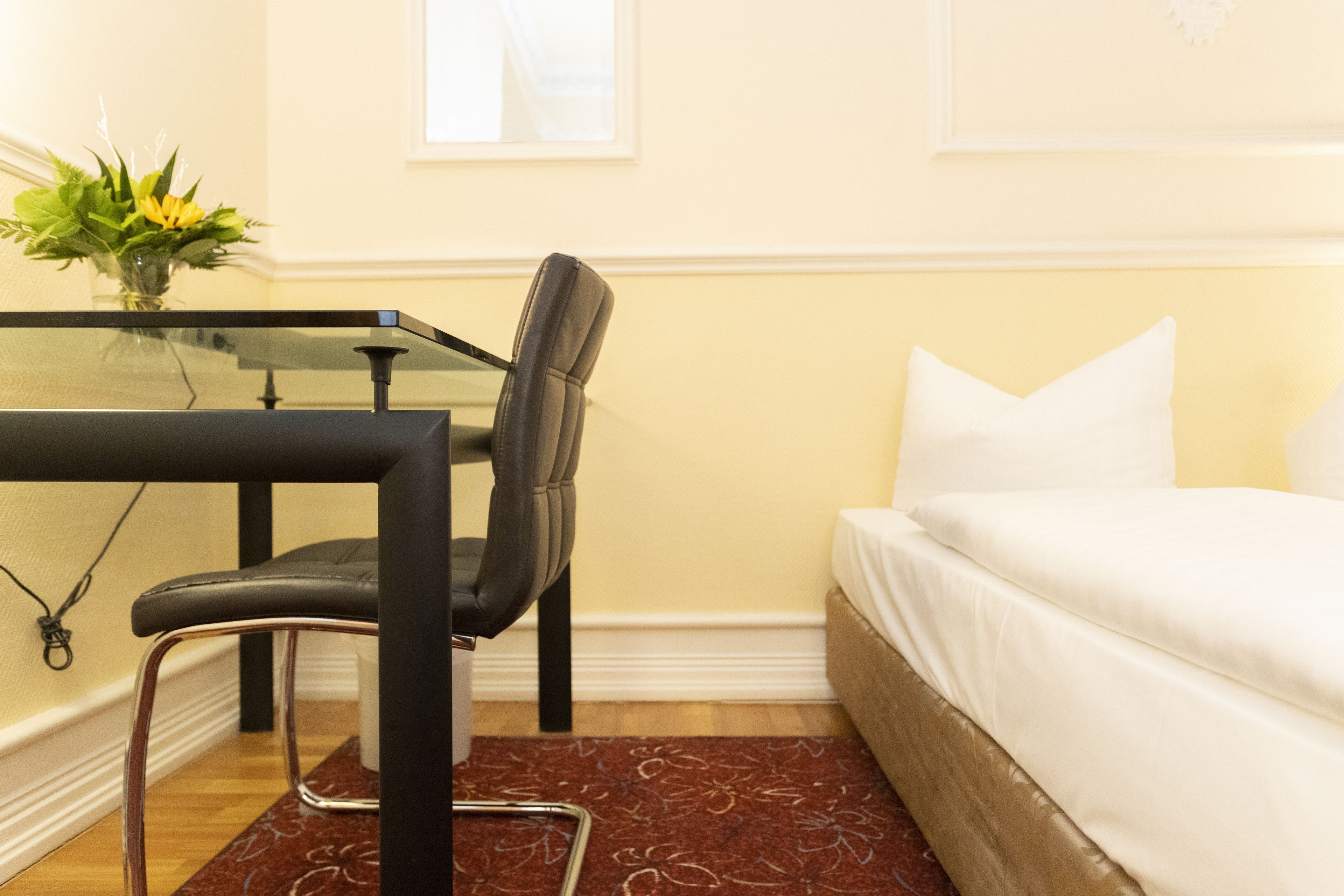 A hotel room with a glass table, a black chair, a white bed, and a red patterned rug.