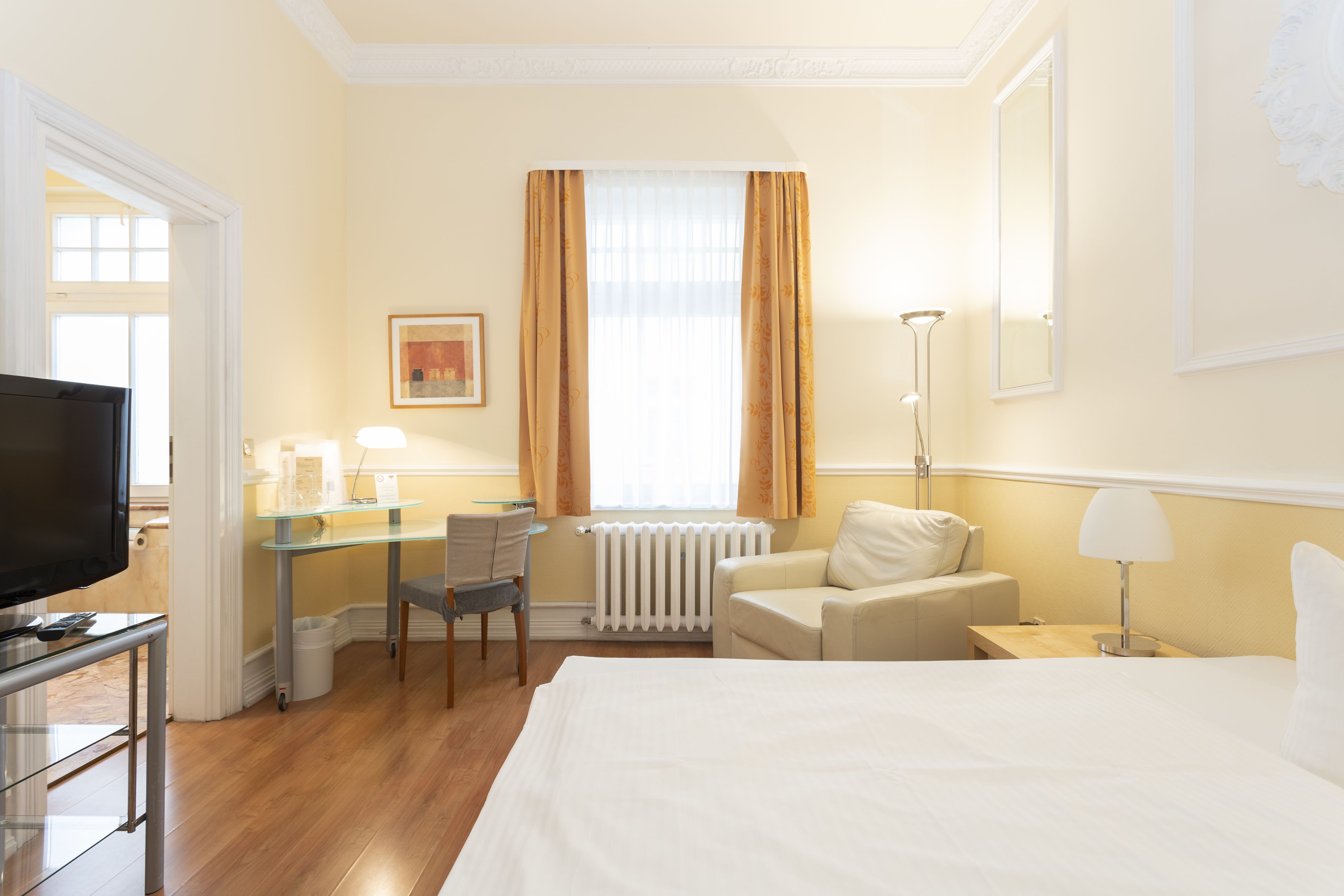 A hotel room with a bed, a desk with a chair, a television, a radiator, and a window with curtains.