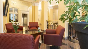 A hotel lobby with a glass coffee table, red upholstered chairs, a large potted plant, and a staircase in the background.