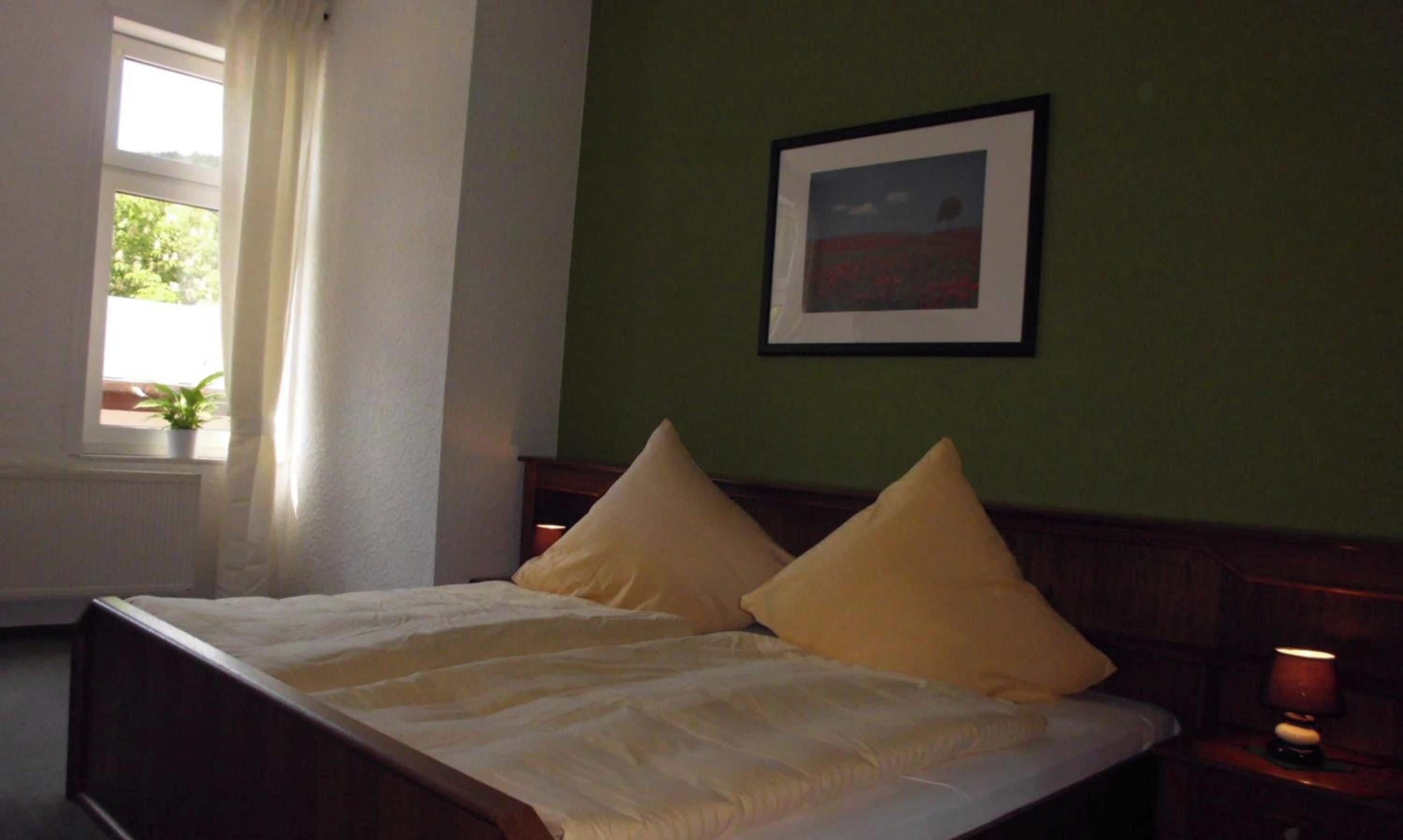 A bed with two pillows, a window with curtains, a framed picture on the wall, and a bedside lamp.