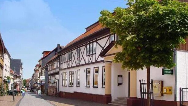 Une rue pavée bordée de bâtiments traditionnels à colombages, un arbre au premier plan et une enseigne indiquant un restaurant.