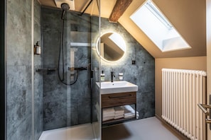 A modern bathroom with a skylight, a glass-enclosed shower, a round mirror, and a white radiator.