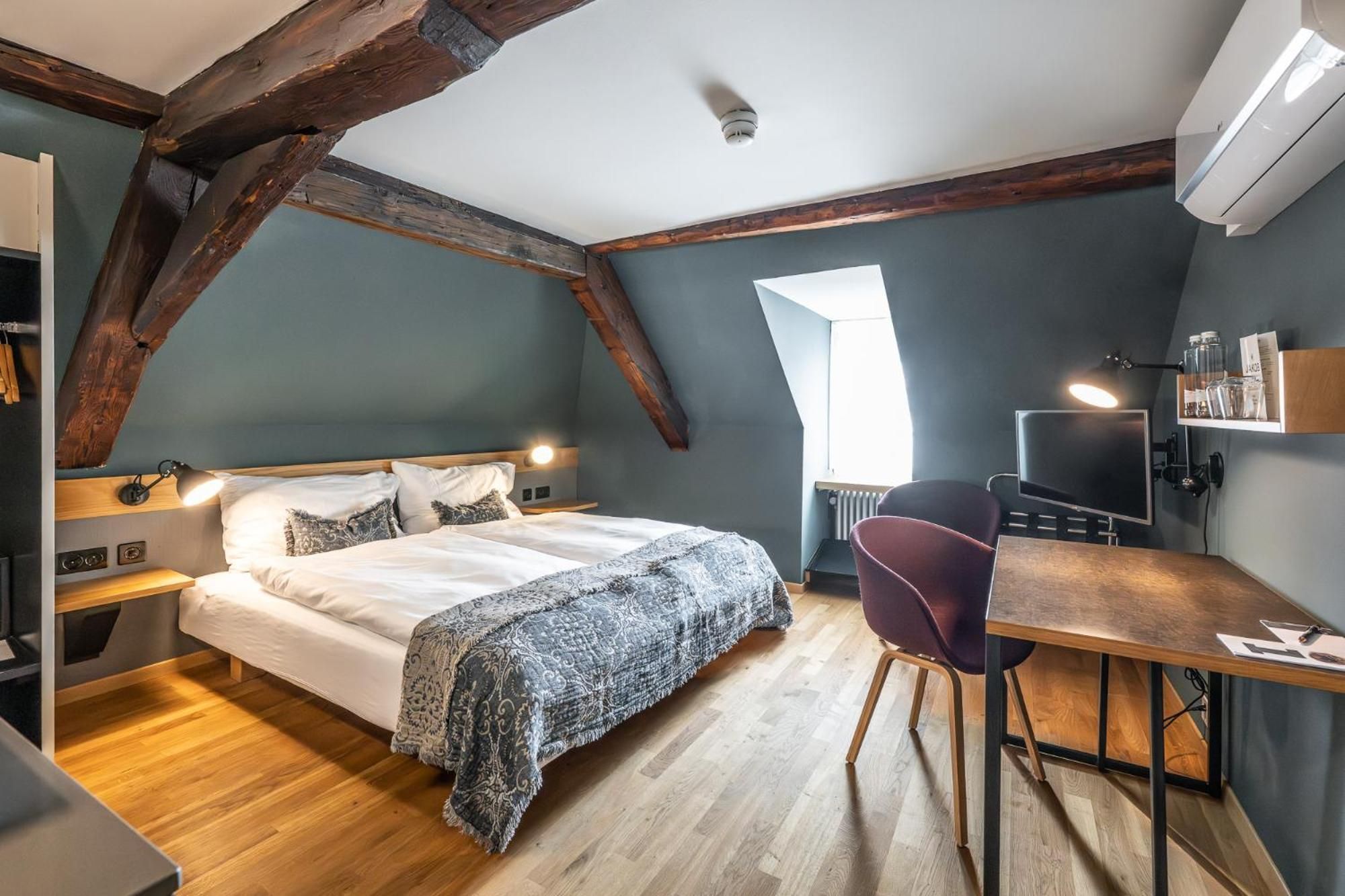 A bedroom with a large bed, a desk, and a chair. There is a window providing natural light.