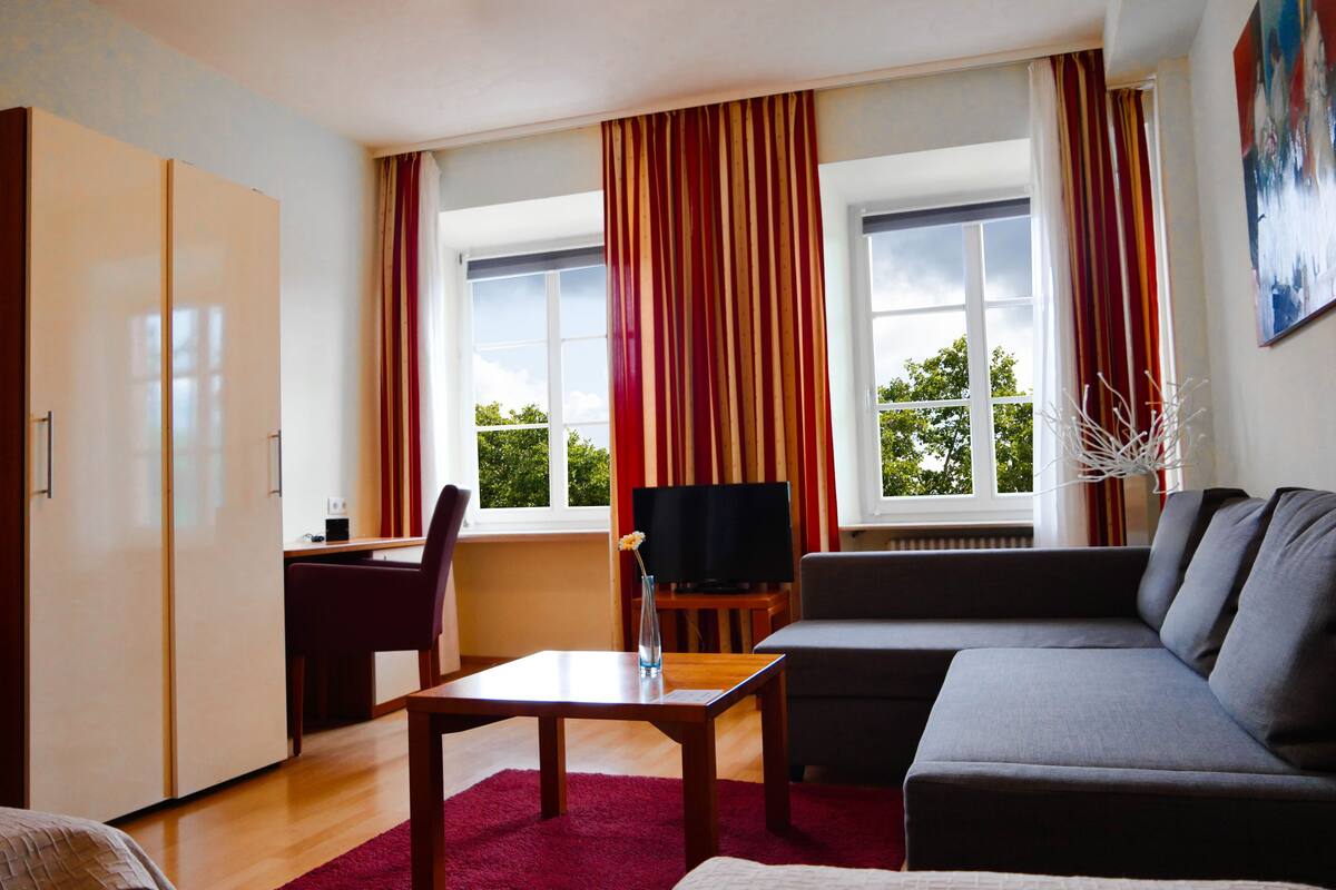 Una sala de estar moderna con un sofá, un televisor sobre un mueble bajo, un escritorio y un ventanal con cortinas.