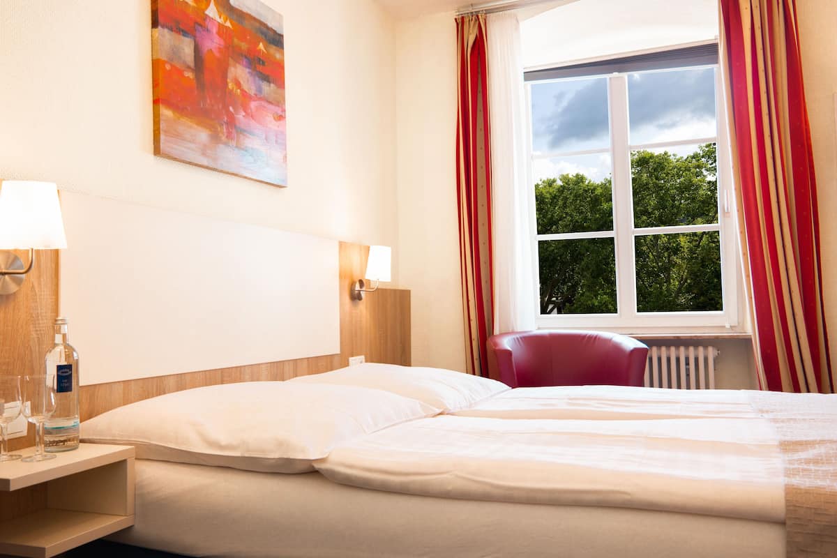 Una habitación de hotel con una cama, una mesita de noche, una lámpara, una botella de agua, una silla roja y una ventana con cortinas.