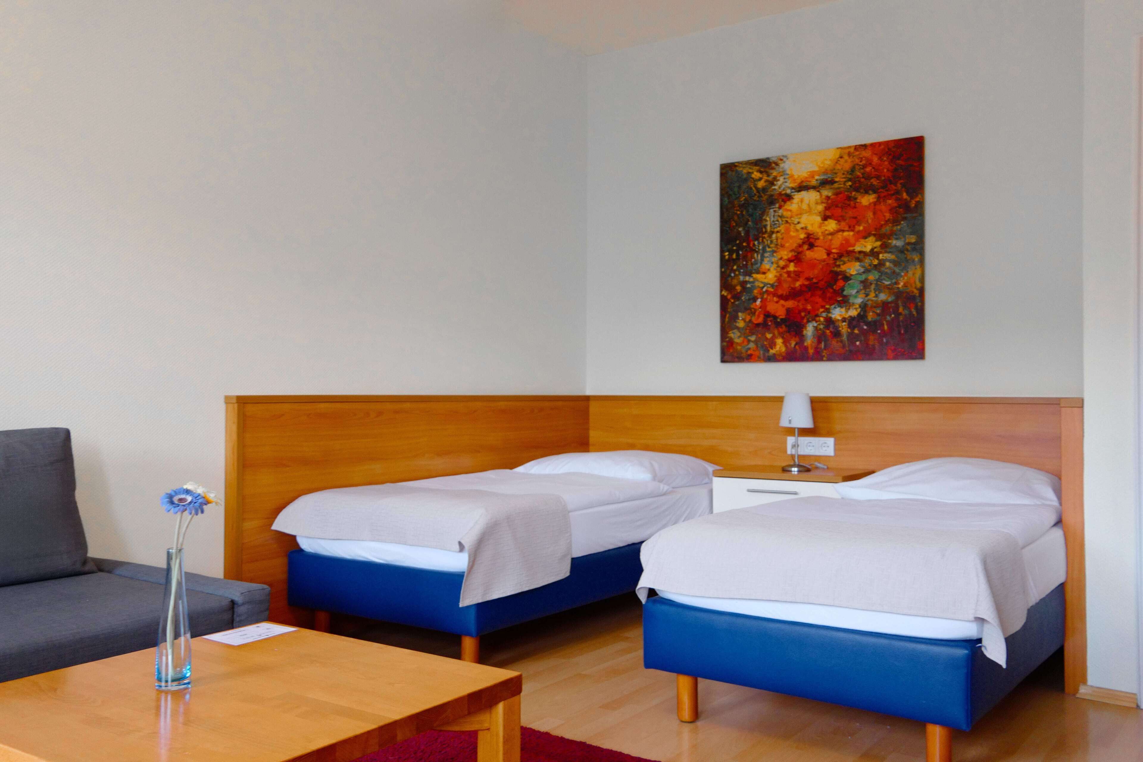 A hotel room with two beds, a wooden headboard, a nightstand with a lamp, a small table, and a vase with flowers.