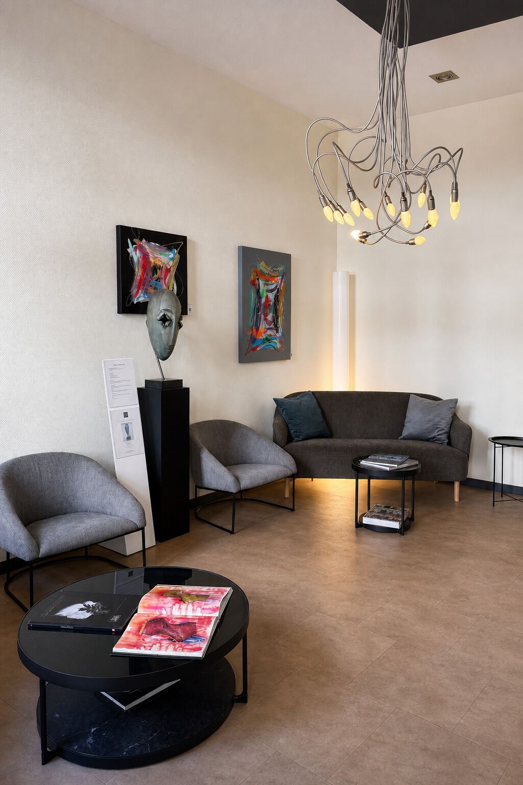 Una sala de estar moderna con un sofá gris, dos sillones, una mesa de centro con revistas, una araña y pinturas abstractas en la pared.