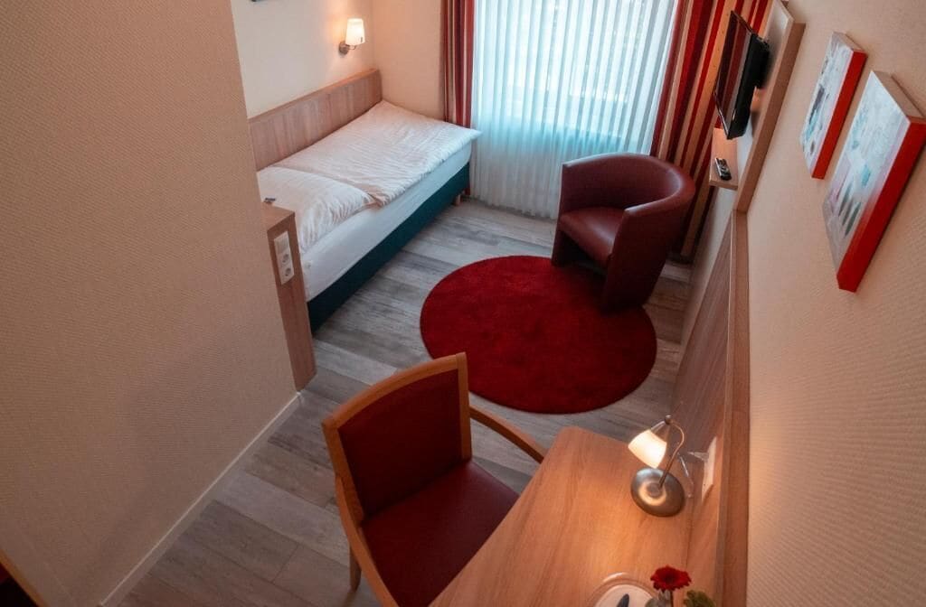 A hotel room with a bed, a red chair, a wooden desk, and a red rug.