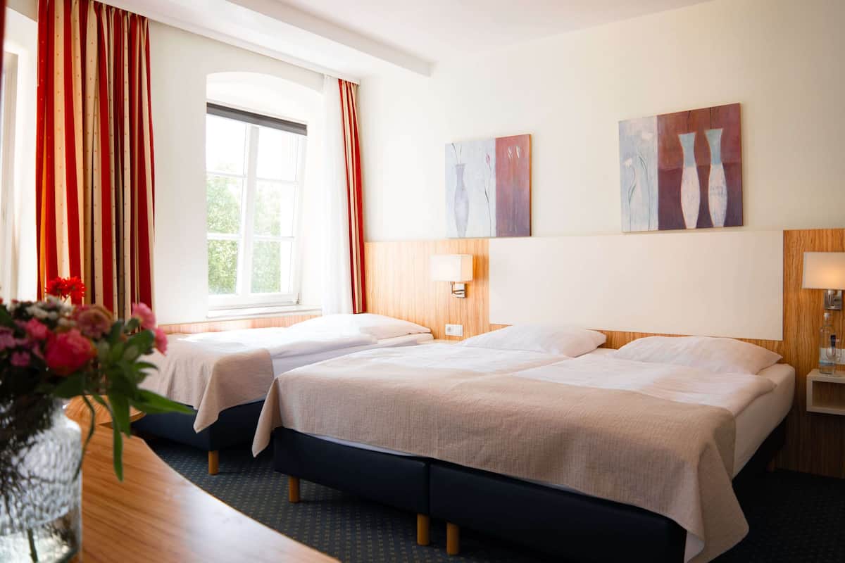 Habitación de hotel con dos camas, un ventanal con cortinas, un jarrón de flores y un cuadro abstracto en la pared.