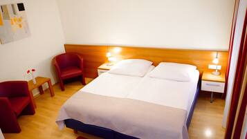 Habitación de hotel con cama doble, dos mesitas de noche con lámparas, un sillón rojo, una pequeña mesa de madera con un florero y una alfombra roja.