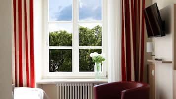 A hotel room with a bed, a red chair, a window with curtains, a radiator, and a TV on the wall.