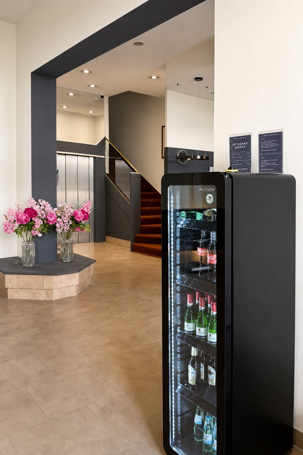 A hotel lobby with a black refrigerator stocked with beverages, a staircase, and a vase with pink flowers on a platform.