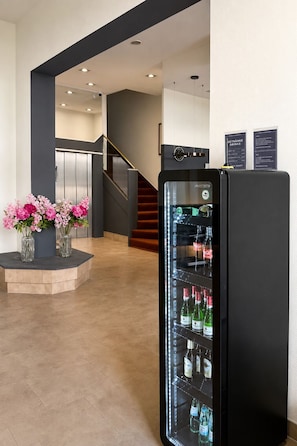 Un vestíbulo de hotel con un refrigerador negro lleno de bebidas, una escalera y un jarrón con flores rosas sobre una plataforma.