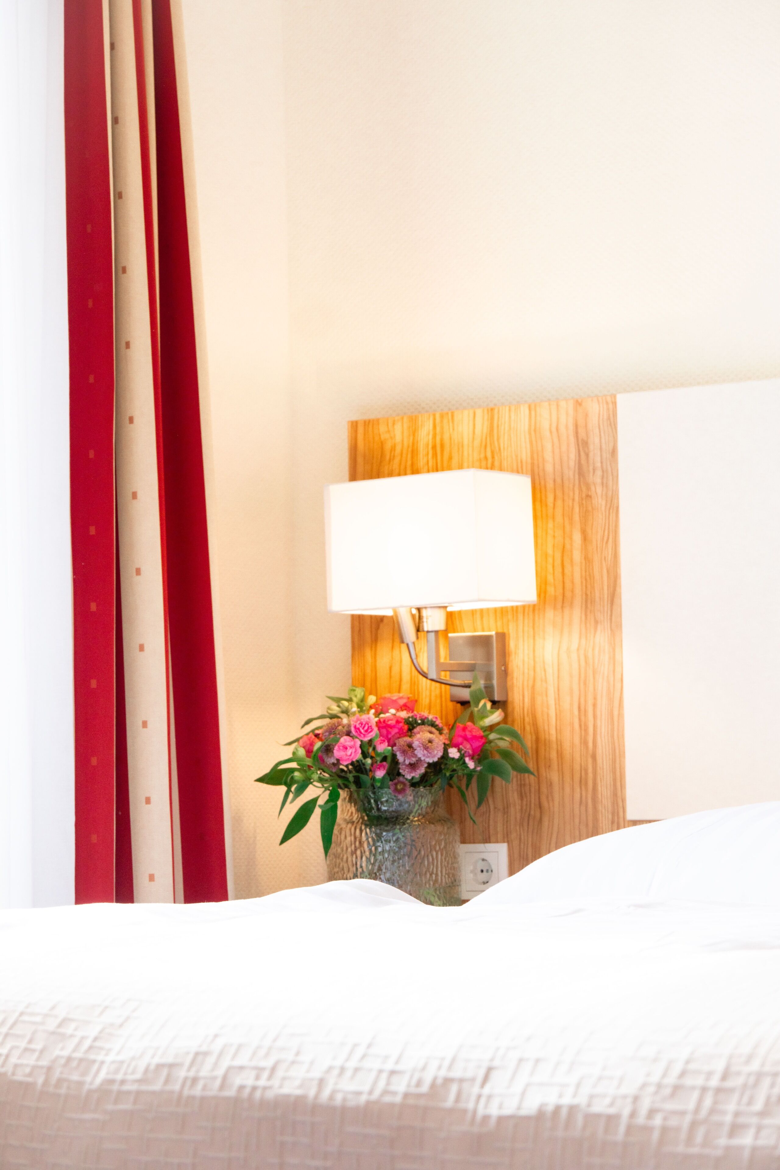 Ein Hotelzimmer mit einem ordentlich bezogenen Bett, einer Nachttischlampe und einer Vase mit Blumen.