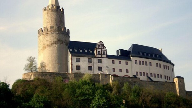 Um castelo histórico com uma torre imponente, um edifício grande com várias janelas e uma muralha ao redor.
