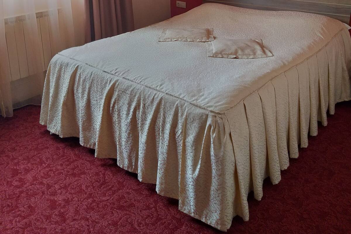 A neatly made bed with a patterned bedspread and pillows, a red wall with a single painting, and a window with sheer curtains.