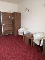 Una habitación con alfombra roja, dos sillones de estampado floral, una mesita de madera y un armario con una lámpara sobre él.