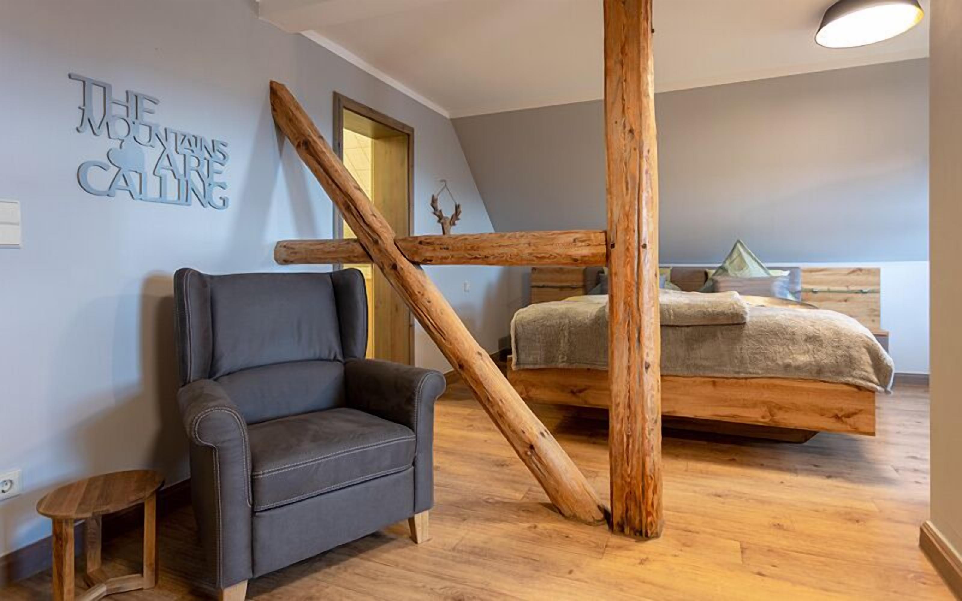 A room with a wooden beam structure, a grey sofa, a bed with a wooden headboard, and a wall sign reading "THE MOUNTAIN CALLING".
