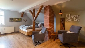 Une chambre de style « loft », avec un plafond en pente, des poutres apparentes, une cheminée en briques, un lit, deux fauteuils et un lampadaire.