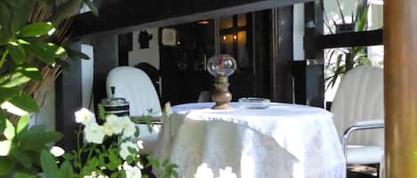 Un área de comedor al aire libre con una mesa redonda cubierta con un mantel blanco, rodeada de follaje y flores blancas.