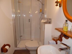 A bathroom with a glass-enclosed shower, a toilet, a sink, and a wooden shelf with toiletries.