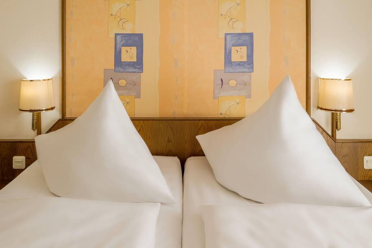 A hotel room with two beds, each with white linens and pillows, flanked by wall-mounted lamps.