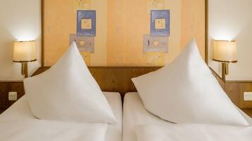 A hotel room with two beds, each with white linens and pillows, flanked by wall-mounted lamps.