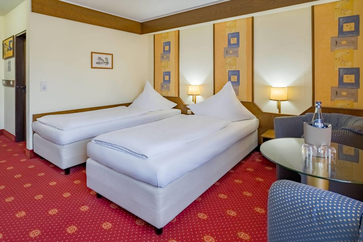A hotel room with two single beds, a small table with glasses and a bottle, and a blue armchair.