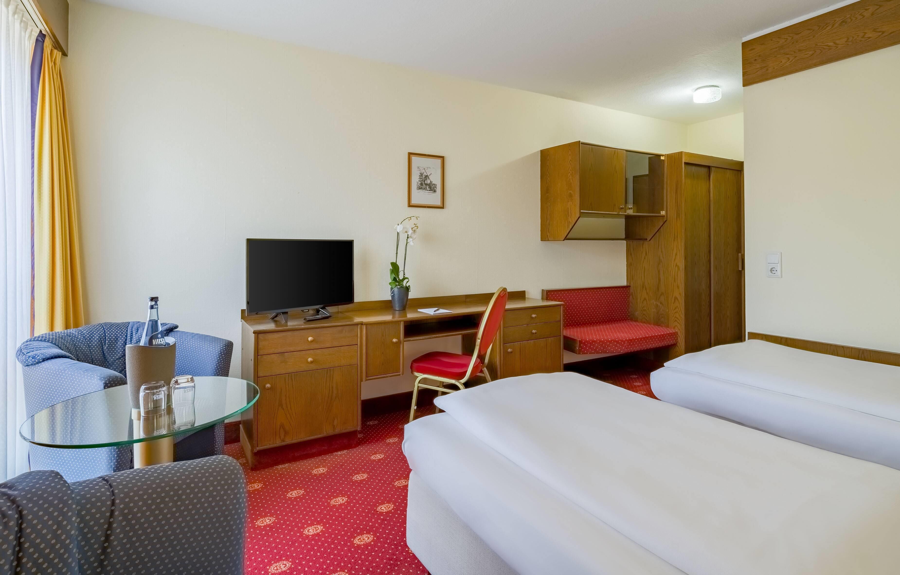 A hotel room with a bed, a desk with a chair, a television, a wardrobe, and a small table with a glass.