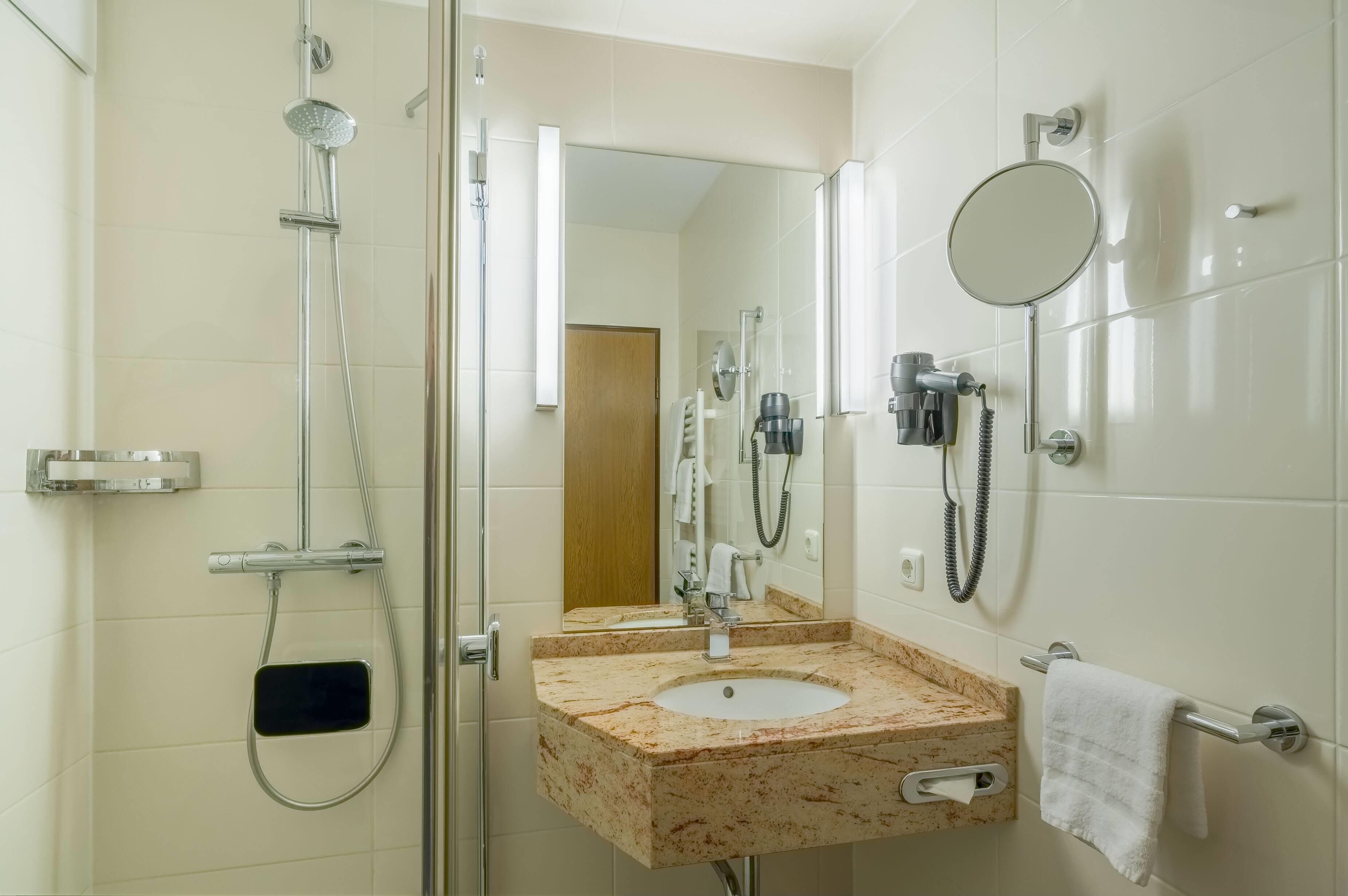 Un baño moderno con ducha envuelta en vidrio, lavamanos de mármol, cabezal de ducha de mano, secador de pelo portátil y un perchero para toallas.