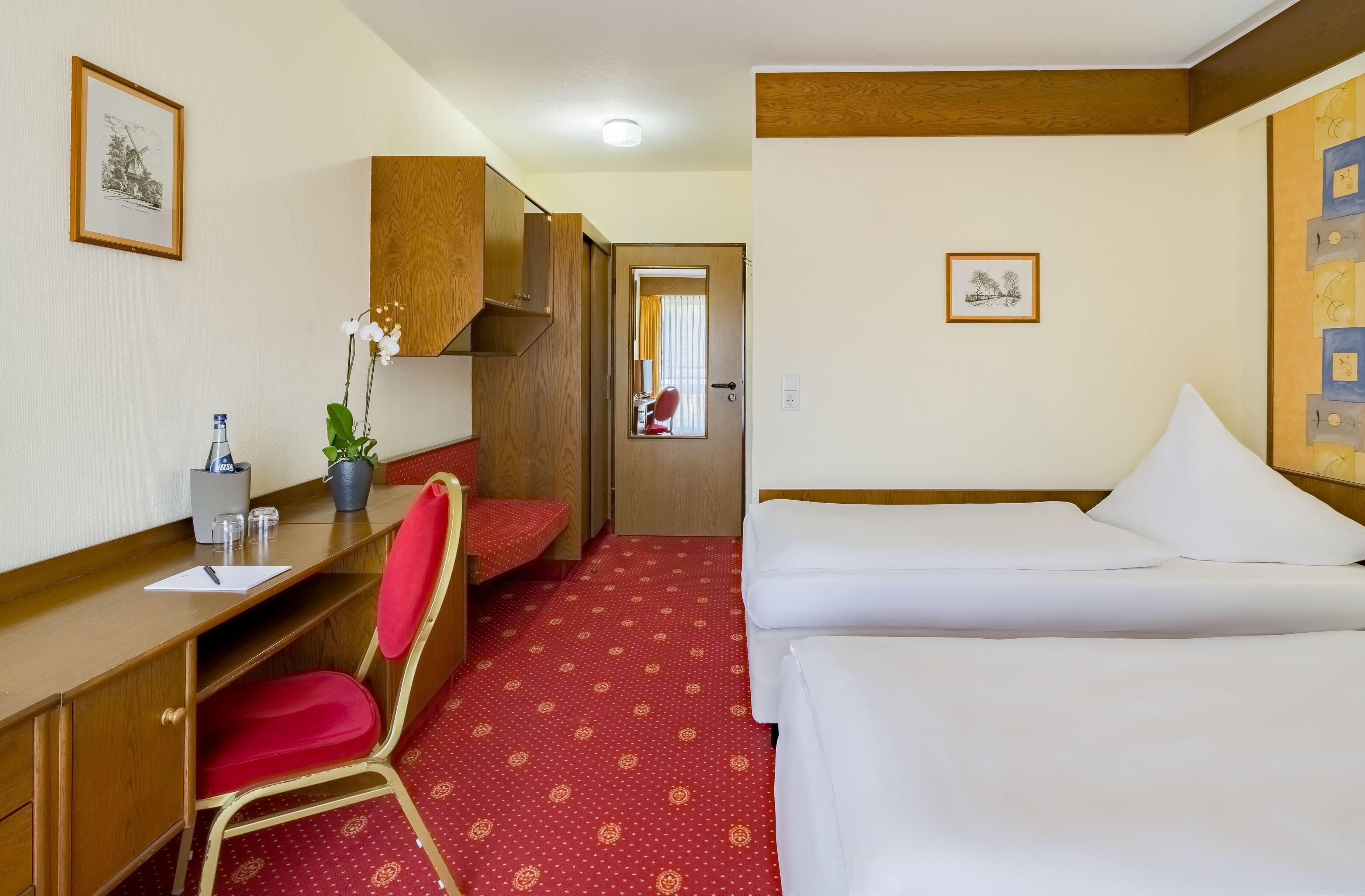 A hotel room with a wooden desk, a red chair, a bed with white linens, and a red patterned carpet.
