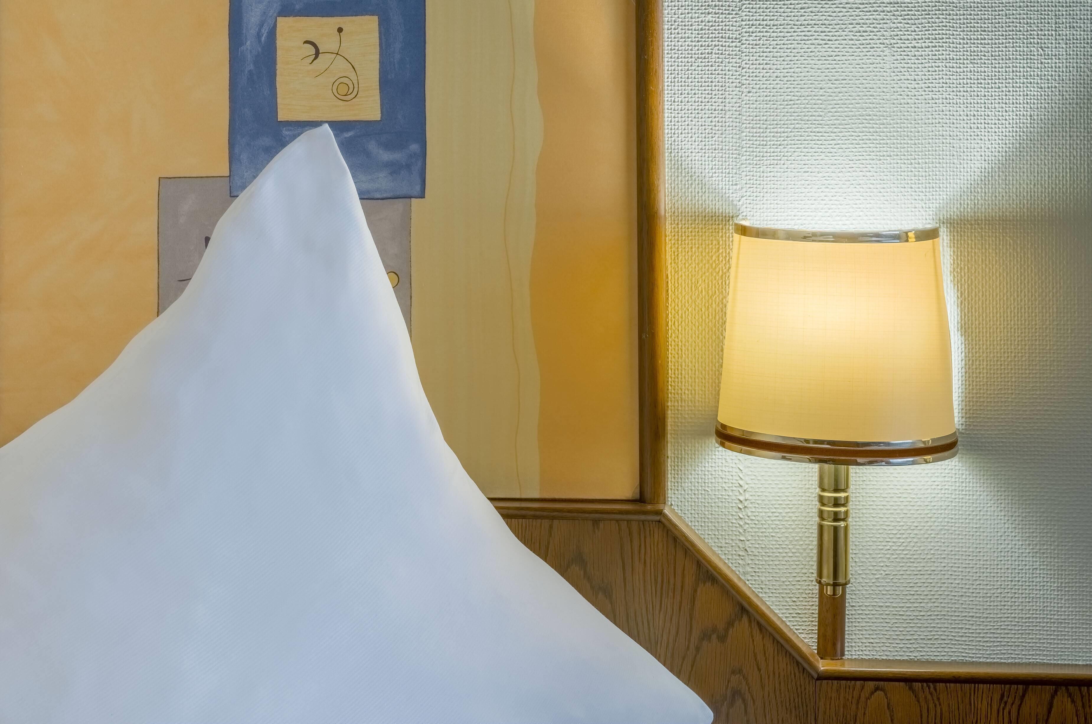 Una habitación de hotel con un almohadón blanco, un reloj de pared y una lámpara con pantalla texturizada.