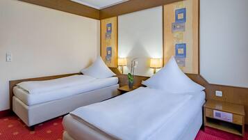 A hotel room with two single beds, a wooden headboard, a small bedside table with a lamp, and a red patterned carpet.