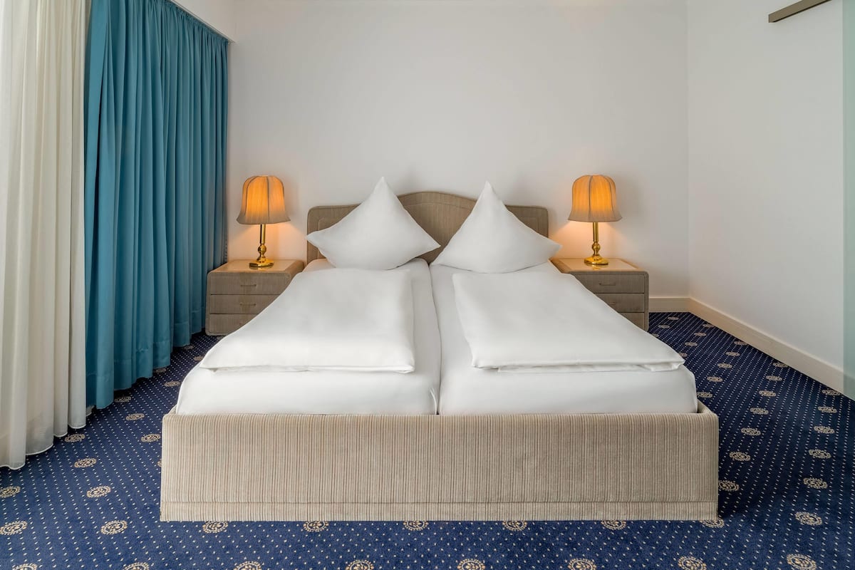 A double bed with white linens and two bedside tables with lamps. The room has a blue carpet with a pattern and a blue curtain on the left.