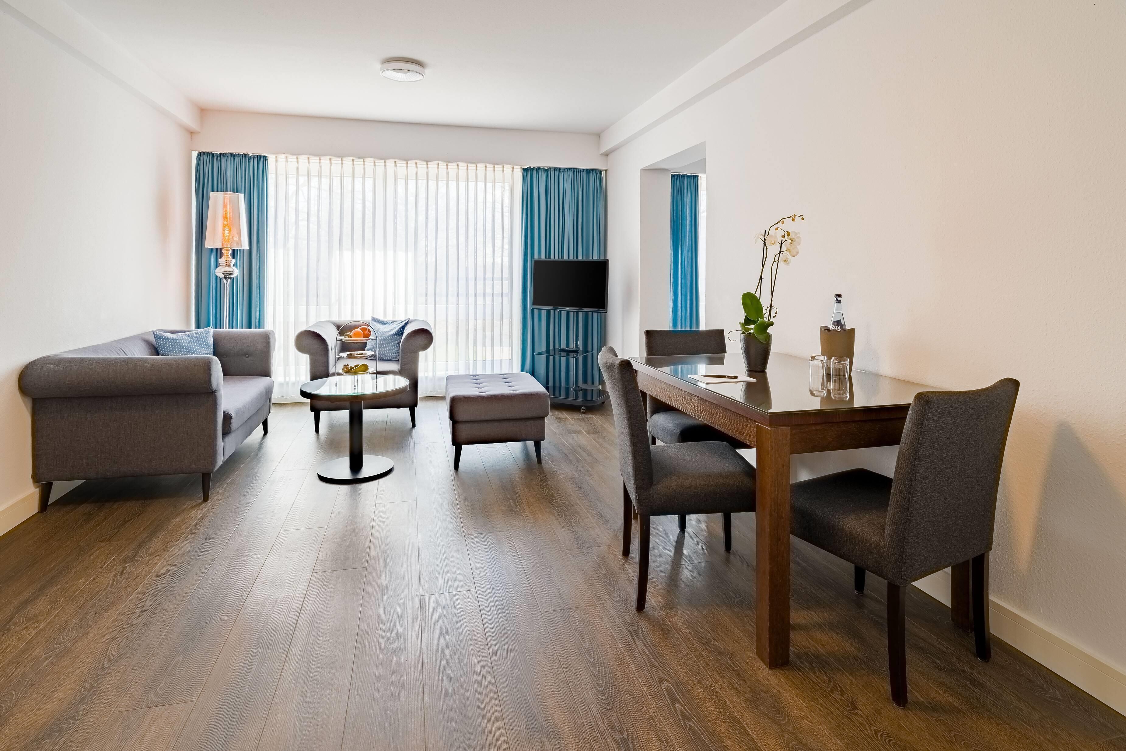 A modern living room with a sofa, armchair, ottoman, coffee table, and dining table with chairs. A flat-screen TV is mounted on the wall, and there is a potted plant on the dining table.