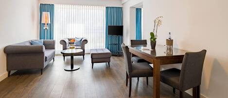 Una sala moderna con sofá, sillón, otomanas, mesa de centro y mesa de comedor con sillas. Un televisor de pantalla plana está montado en la pared, y hay una planta en maceta sobre la mesa del comedor.