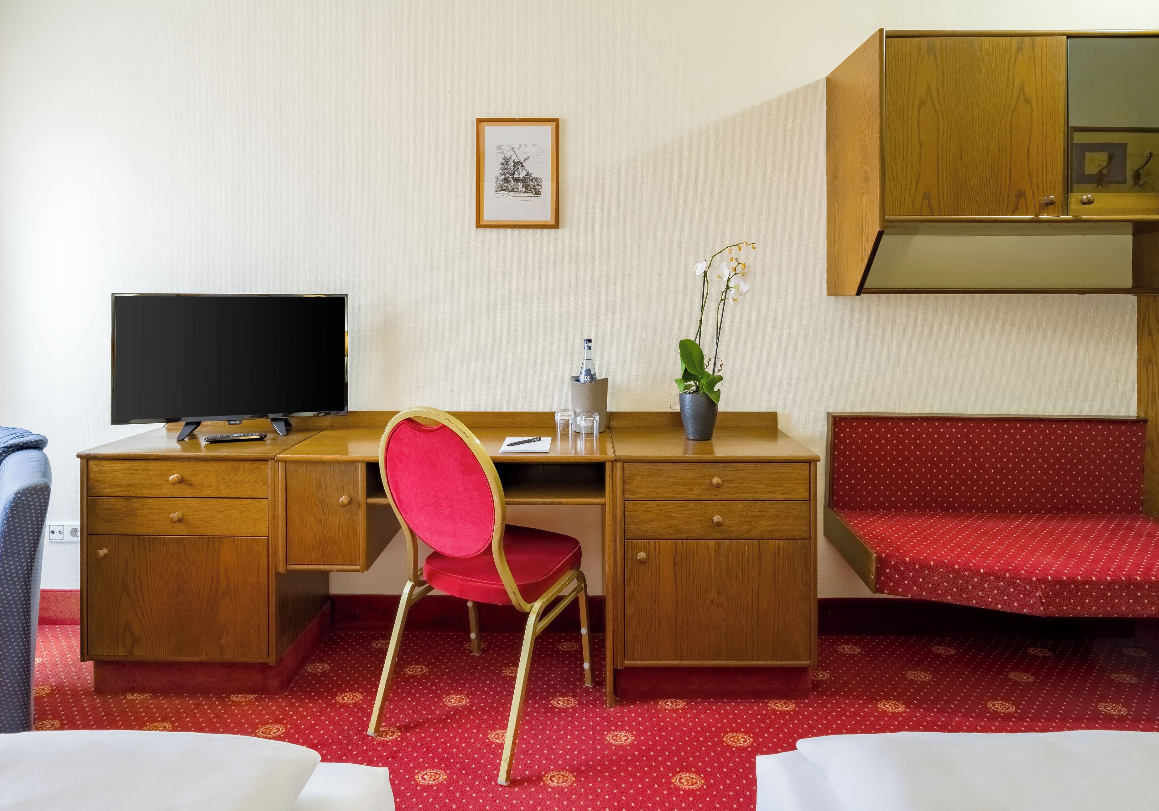 Habitación de hotel con un escritorio de madera, una silla roja, un televisor, una planta pequeña y un sofá rojo.