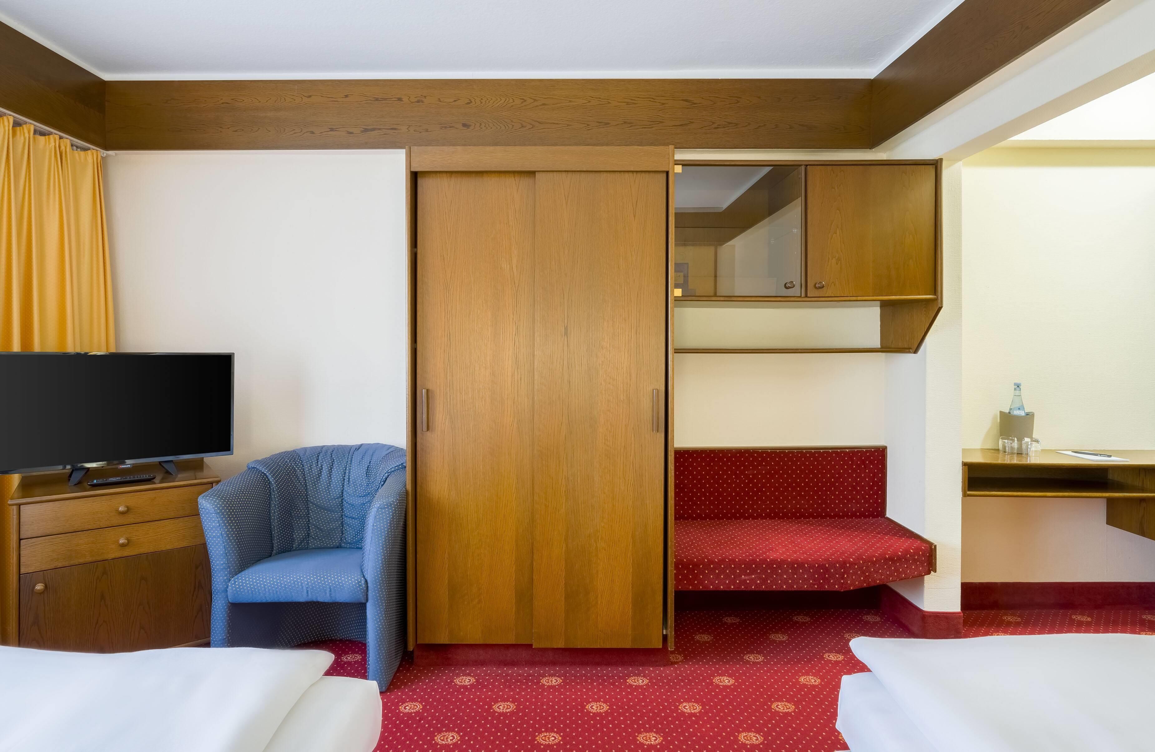 A hotel room with a TV, a blue chair, a wardrobe, a desk with a mirror, and a bed.
