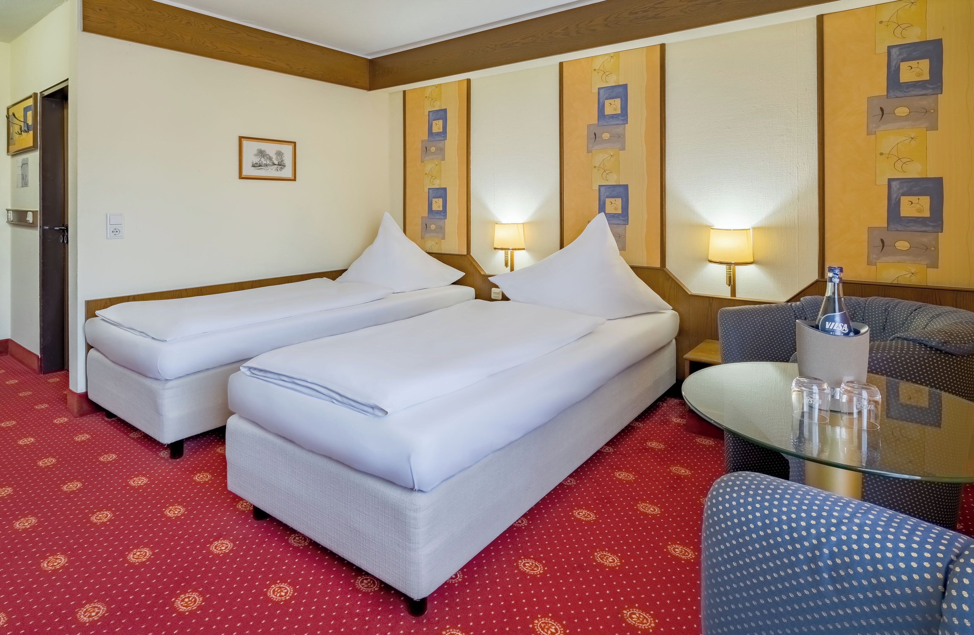 A hotel room with two single beds, a small table with glasses, a chair, and a wall with framed pictures.