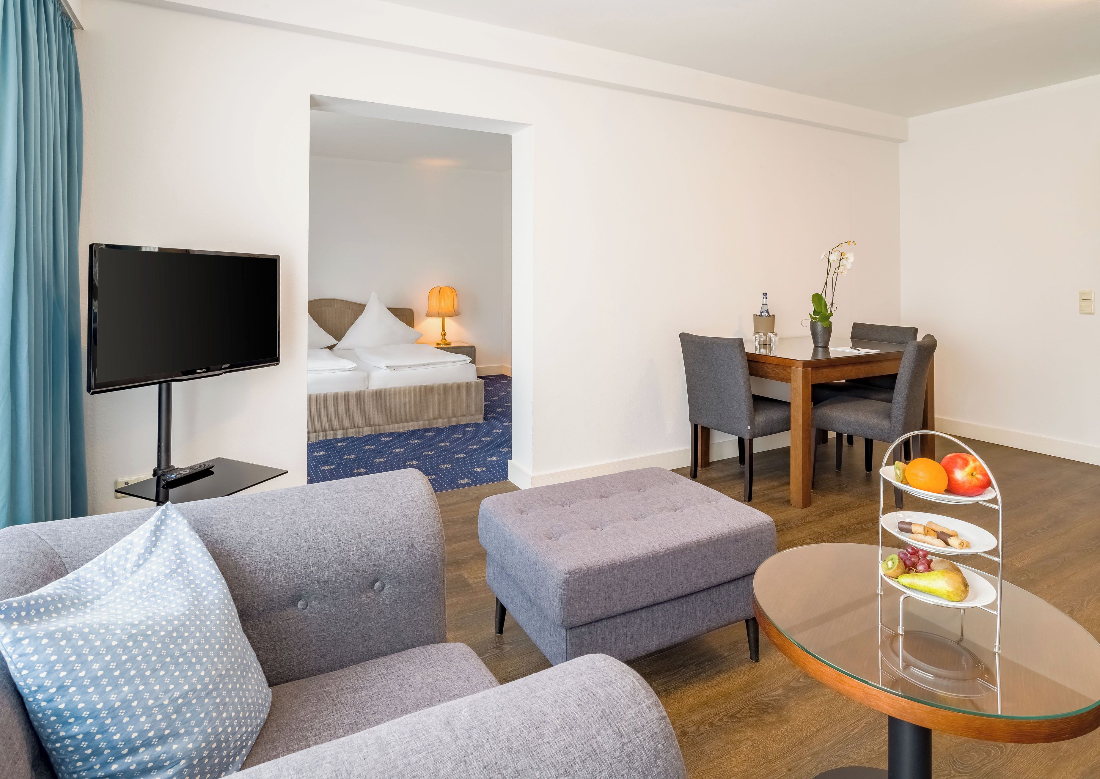 Habitación de hotel con televisor, sofá, reposapiés, mesa de comedor, sillas y una pequeña mesa con fruta.