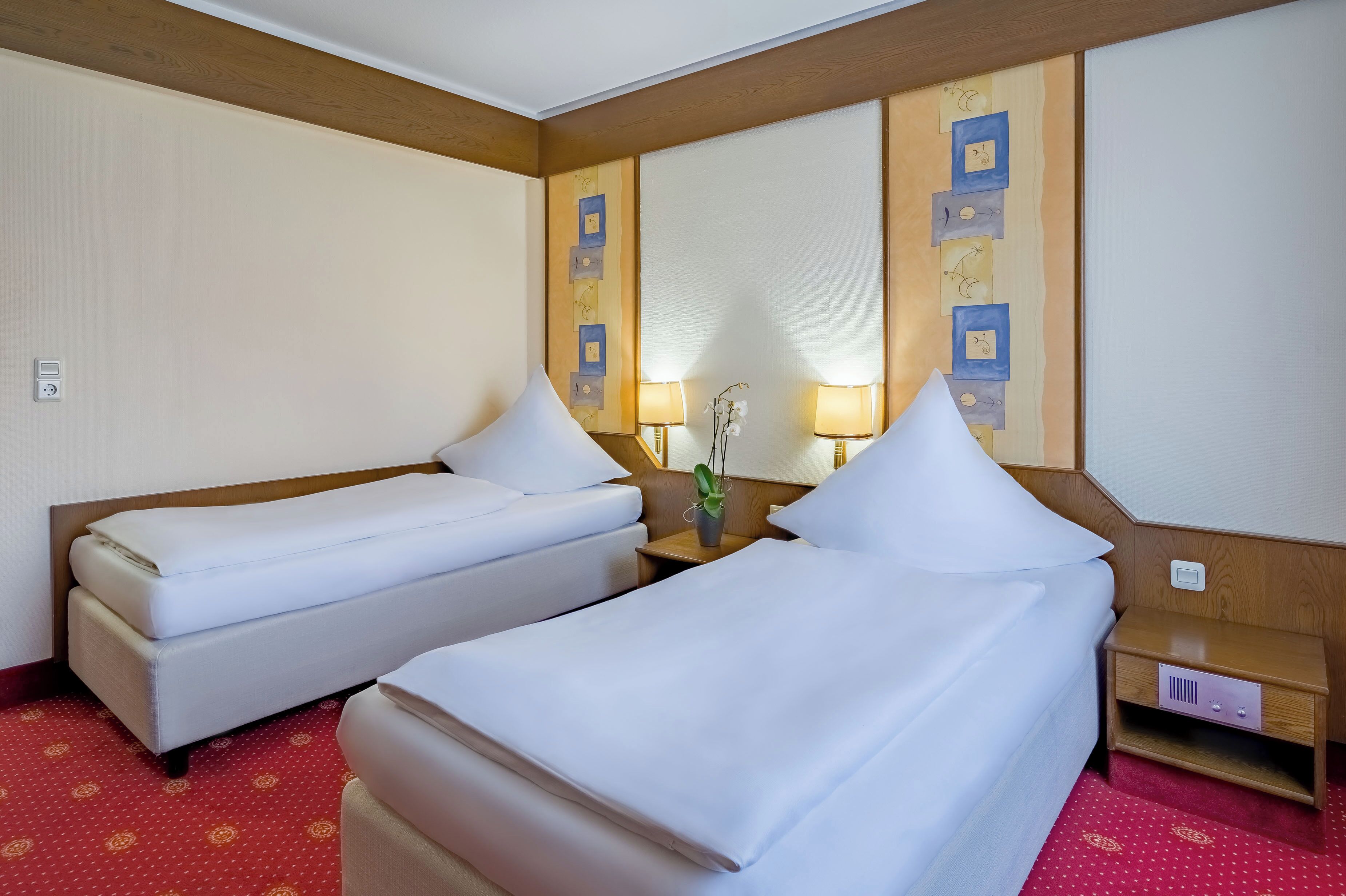A hotel room with two single beds, a wooden headboard, a small bedside table with a lamp, and a wall-mounted artwork.