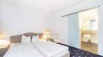 Une chambre d’hôtel avec un lit, des tables de chevet, une lampe et une salle de bain visible à travers une porte ouverte.