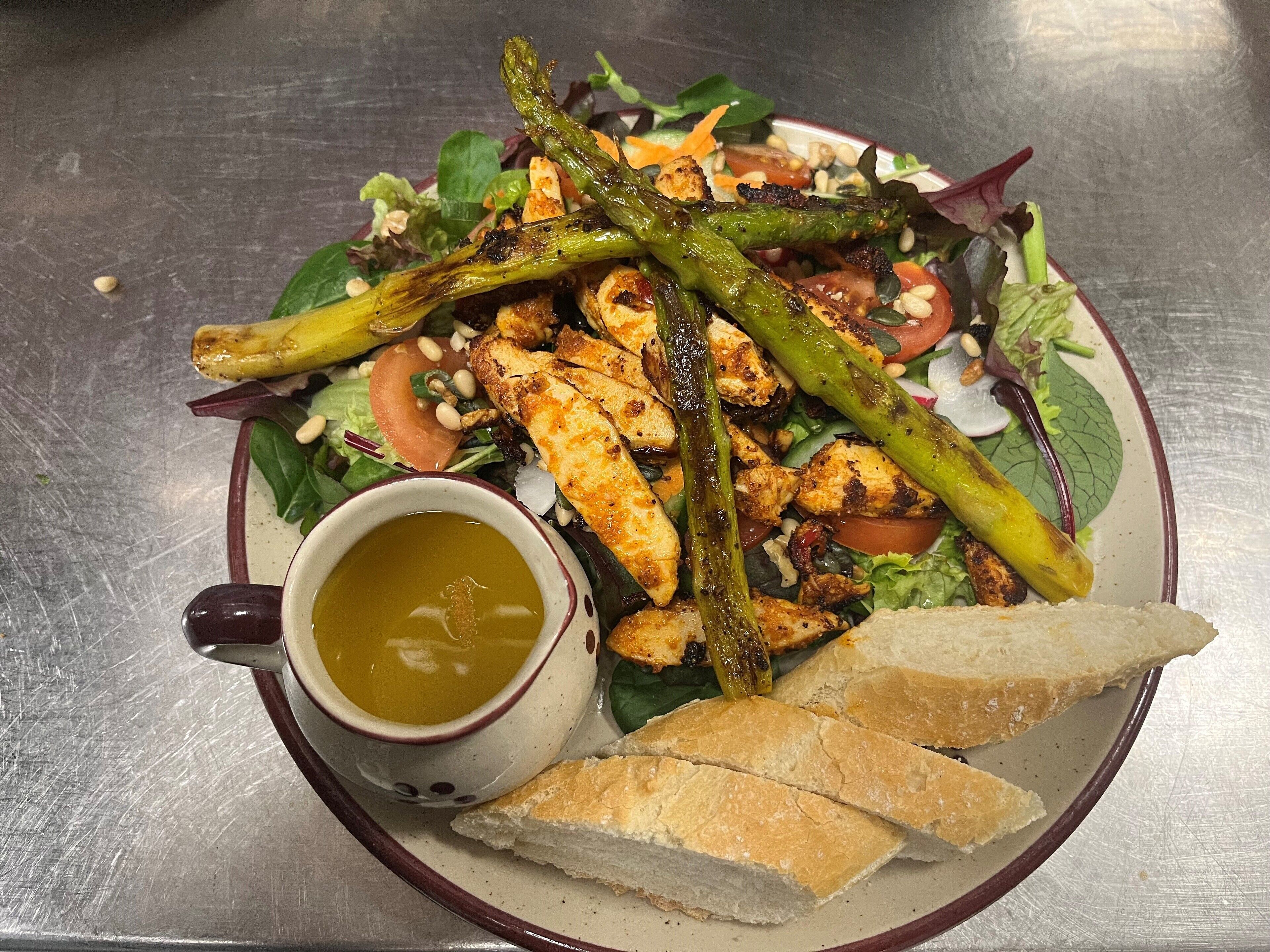 Une assiette de poulet grillé, d’asperges et de salade, accompagnée d’une portion de pain et d’un bol de soupe.