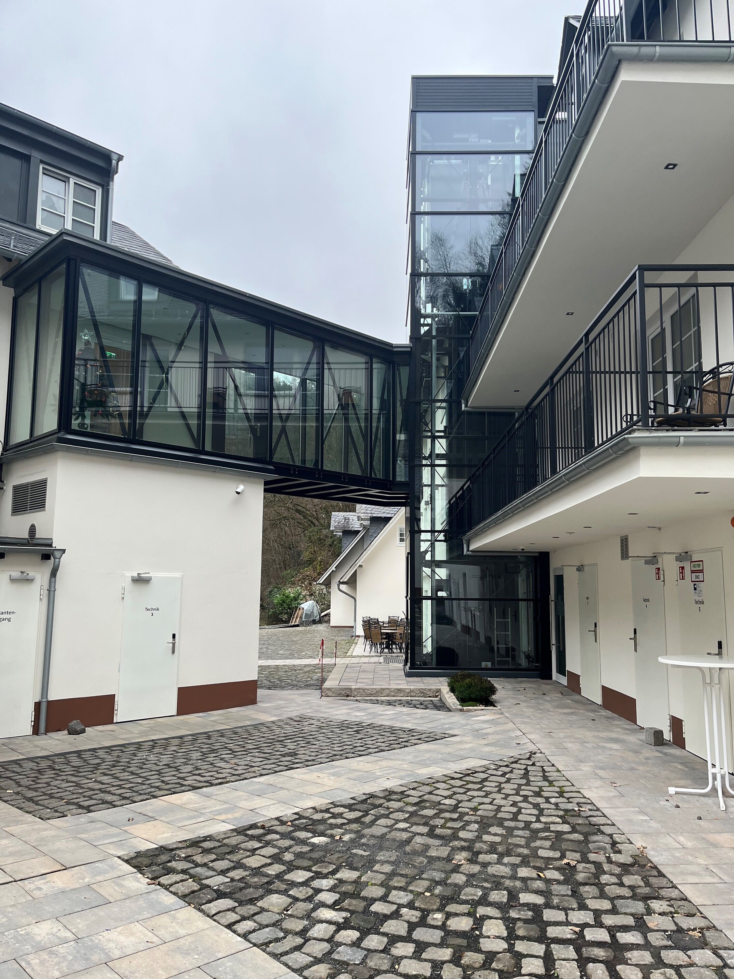 A modern building with a glass bridge connecting two sections, outdoor seating, and a cobblestone courtyard.