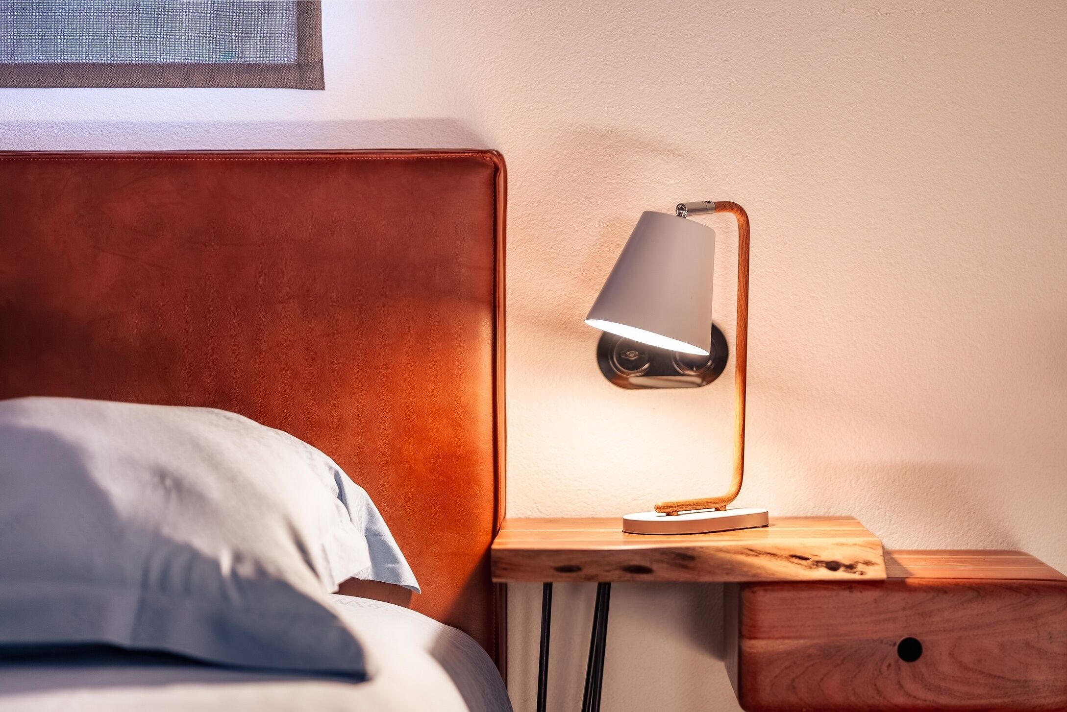 Una cama bien hecha con un almohadón, una mesita de noche de madera con una lámpara y un cajón pequeño.