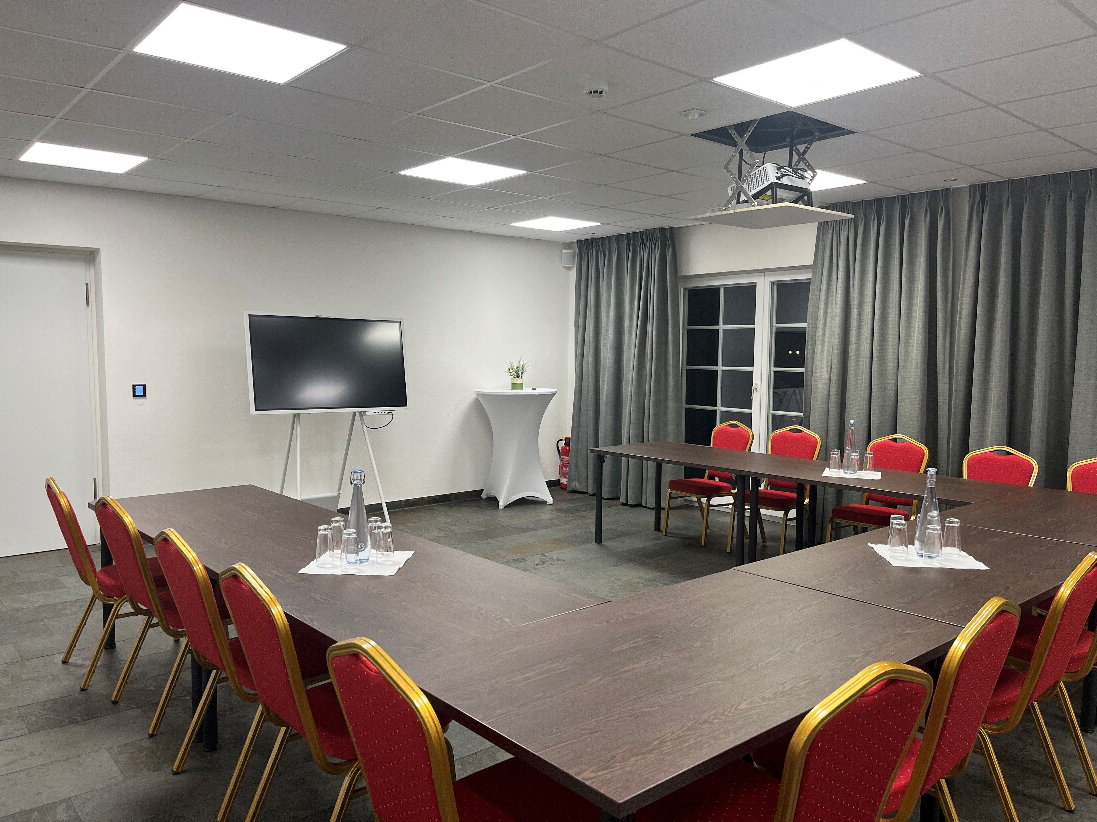Un salón de conferencias con una larga mesa, sillas rojas, un televisor de pantalla plana, un proyector y una ventana con cortinas.